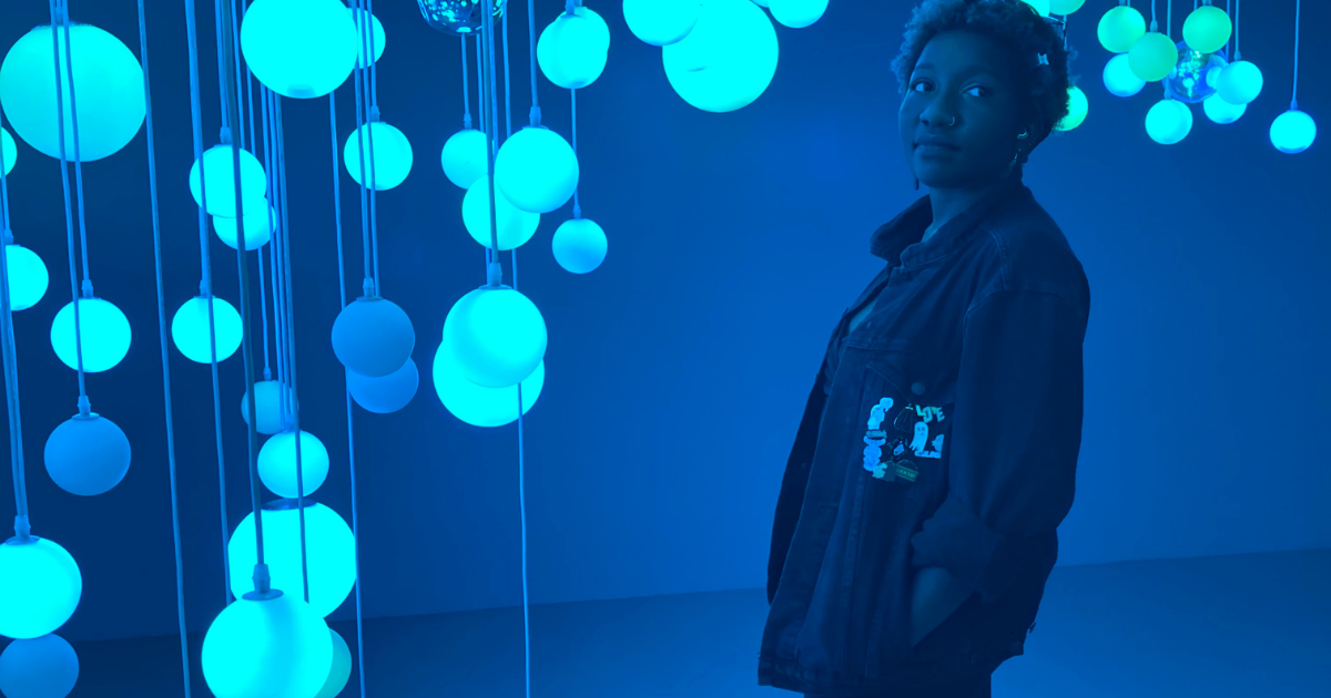 Fati standing in a green blue room with bright teal lights hanging from the ceiling