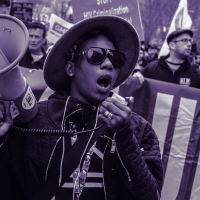 Person holding a megaphone in front of a number of marchers with picket signs that say Silent=Death on the street in front of tall buildings. Purple and grey duotone.
